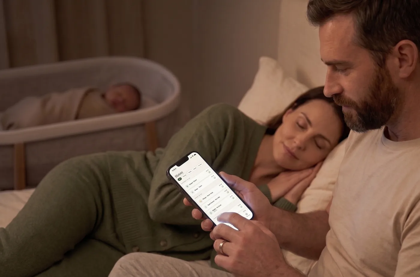 A father checking the shared Little Bean log at night while his partner sleeps beside him and their newborn sleeps in a bassinet